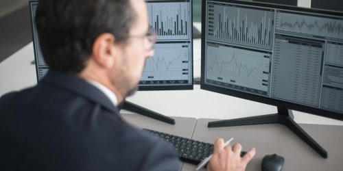 A businessman in a suit analyzing financial charts and graphs on two computer monitors indoors.